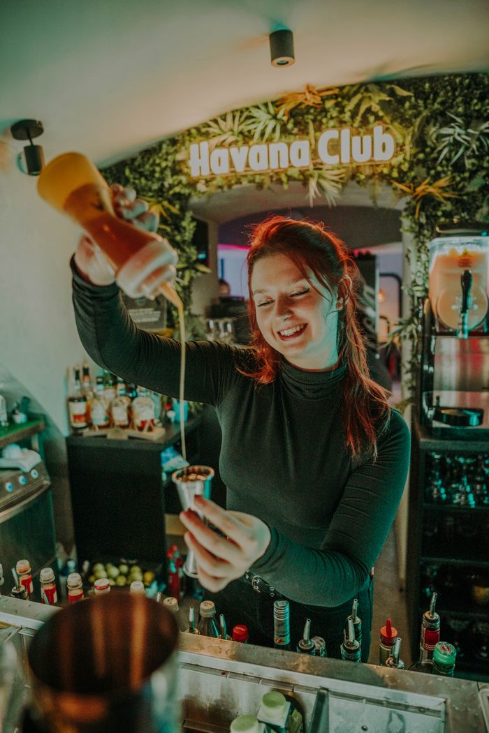 A cheerful bartender skillfully pours a cocktail in a lively Havana Club bar setting, surrounded by various bottles.