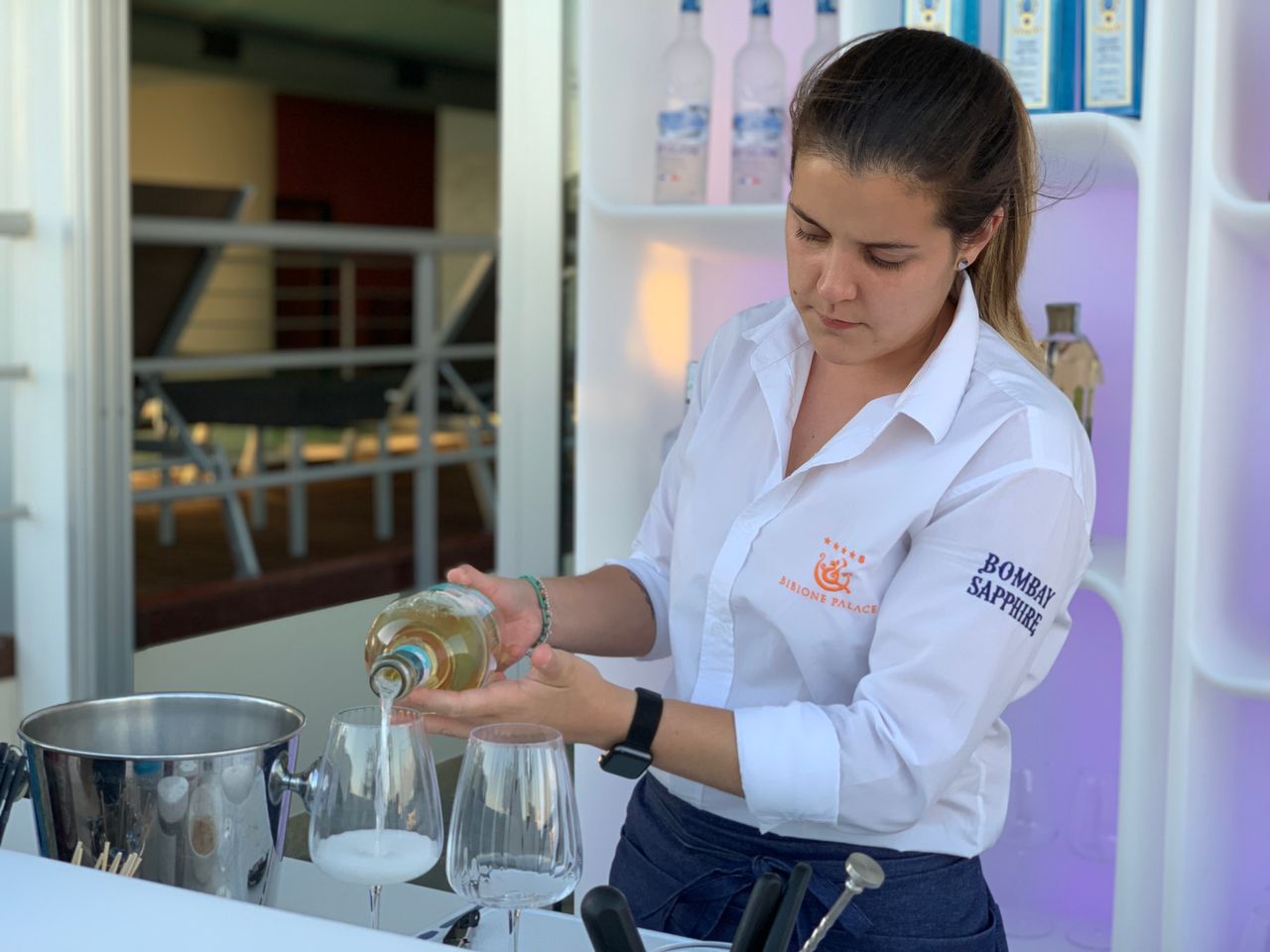 A bartender in a white uniform pouring drinks at an outdoor bar with bottles in the background.