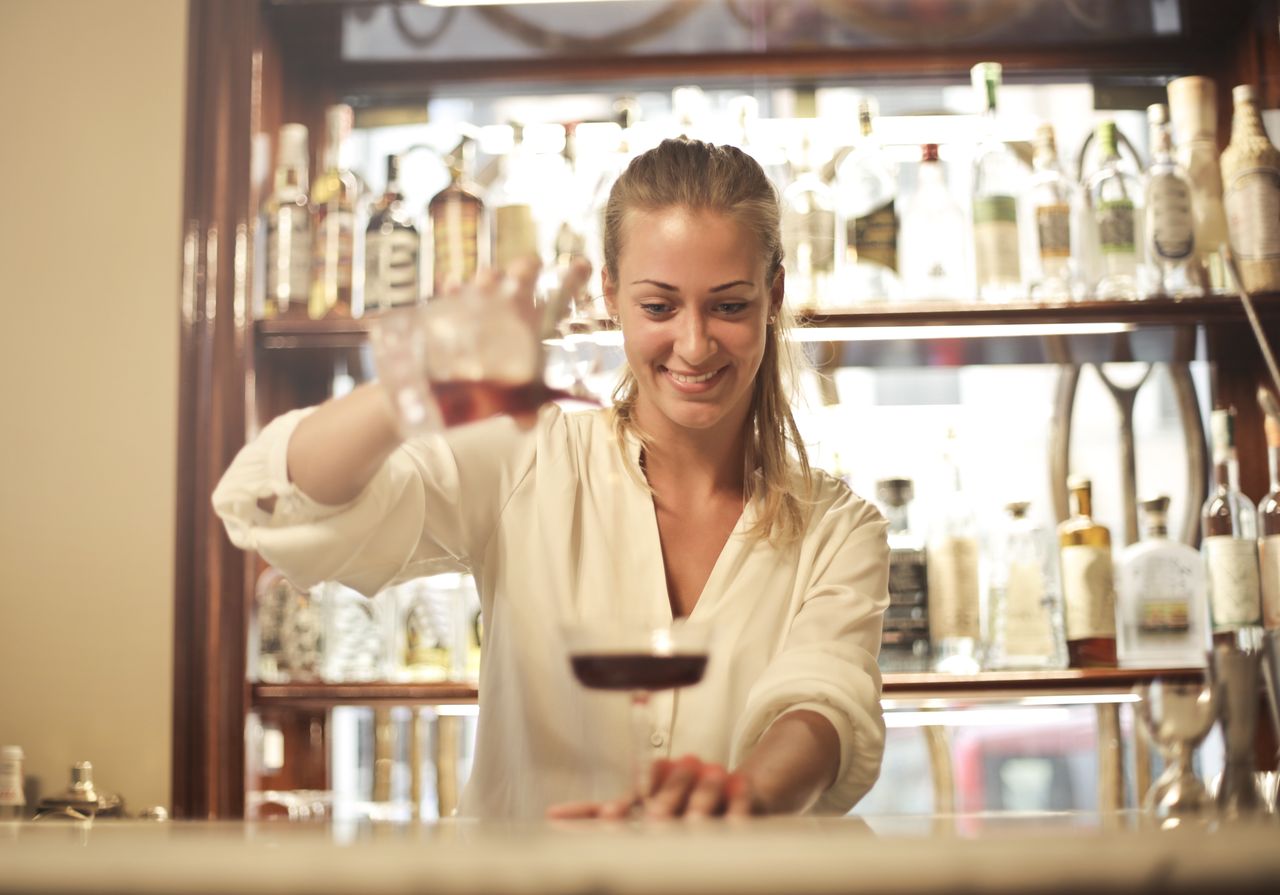 Smiling barmaid in casual clothes standing at counter and pouring alcoholic beverage into glass while making cocktail